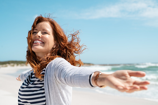 Happy mature woman with arms outstretched feeling the breeze at beach. Beautiful middle aged woman with red hair and arms up dancing on beach in summer during holiday. Mid lady in casual feeling good and enjoying freedom with open hands at sea, copy 