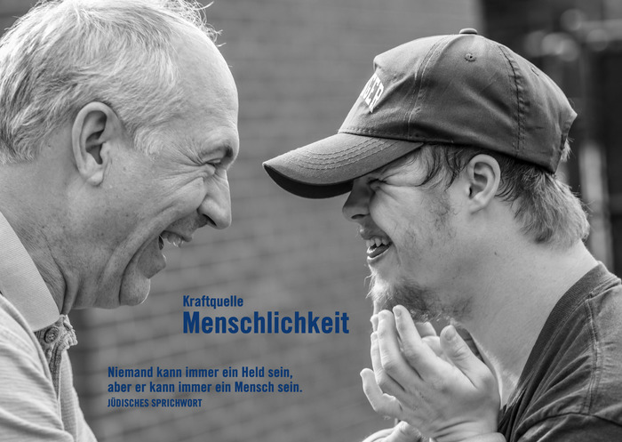 Ein älterer Mann und ein Kind mit Down-Syndrum stehen sich gegenüber und lachen sich an