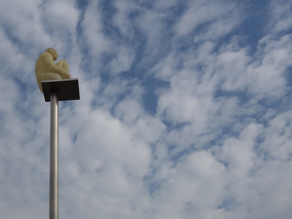 Skulptur: eine Person sitzt auf einer in den Himmel ragenden Säule          