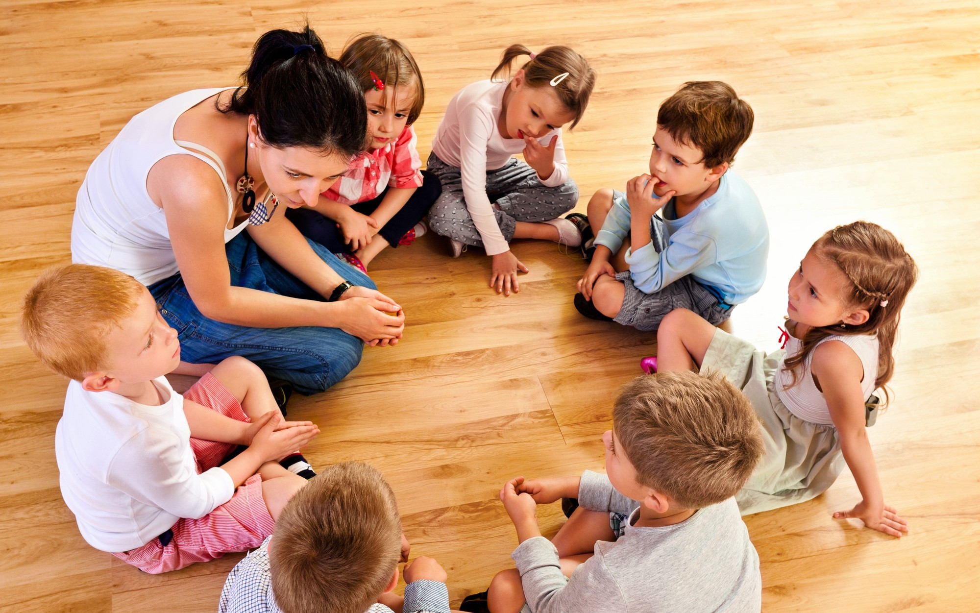 Nursery school children sitting in circle on the floor in a playroom and listening to their teacher. High angle view.
