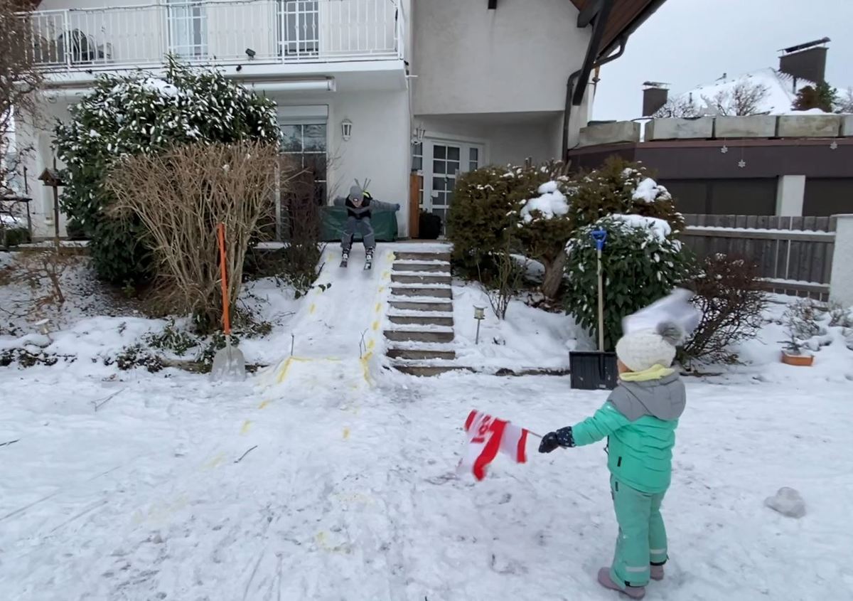 Kind fährt mi Skiern auf Skisprungschanze im Garten zu, während die Tochter mit einer Flagge in der Hand das Startzeichen gibt.