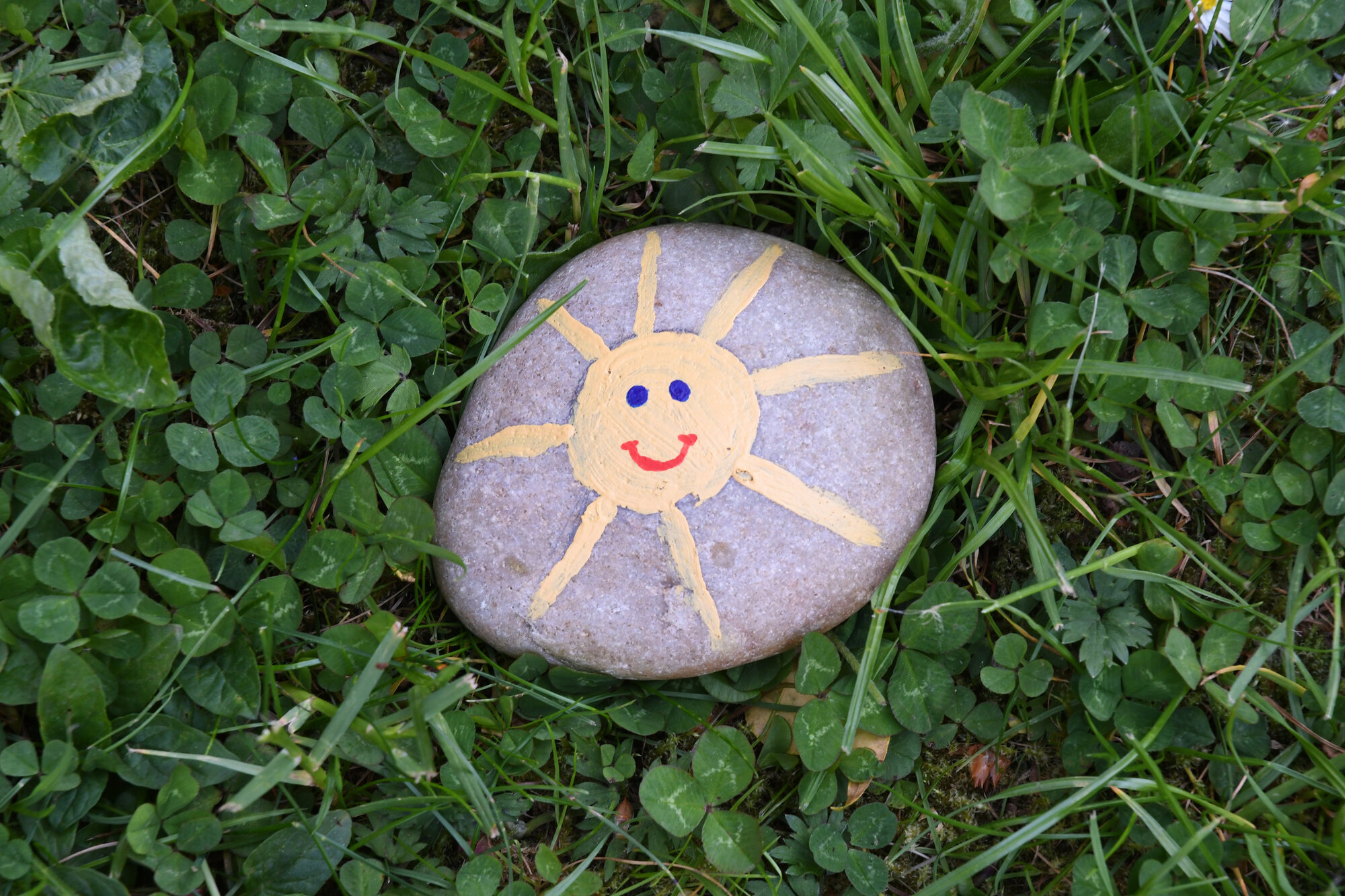 Bemalter Stein mit gelber Sonne und Gesicht, auf grünem Gras liegend.