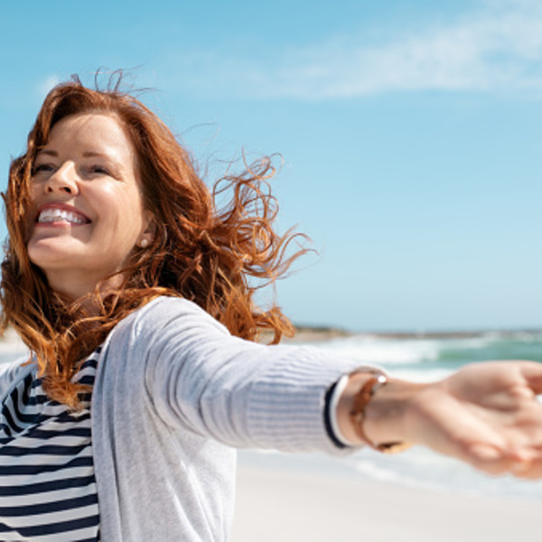 Happy mature woman with arms outstretched feeling the breeze at beach. Beautiful middle aged woman with red hair and arms up dancing on beach in summer during holiday. Mid lady in casual feeling good and enjoying freedom with open hands at sea, copy 