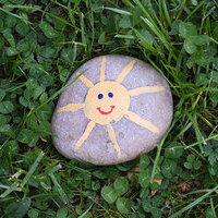 Bemalter Stein mit gelber Sonne und Gesicht, auf grünem Gras liegend.