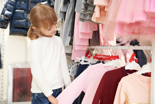Fashion children clothing on hangers in rows at department store. Young, beautiful, pretty girl looking and choosing pink blouse in stylish boutique. Children vogue and style.