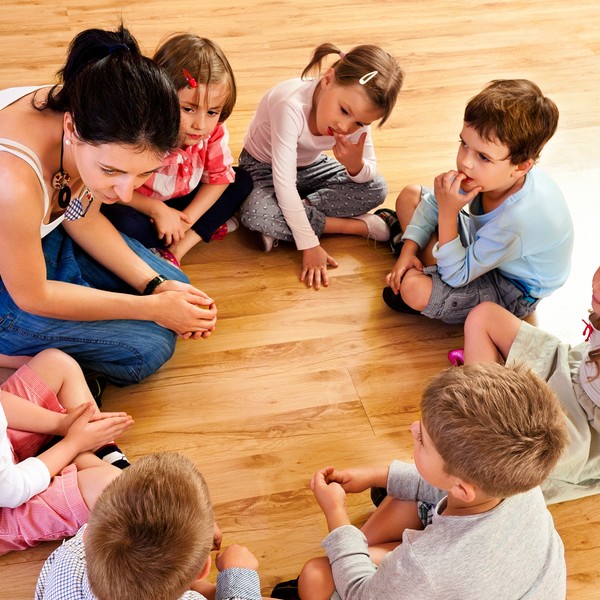 Nursery school children sitting in circle on the floor in a playroom and listening to their teacher. High angle view.