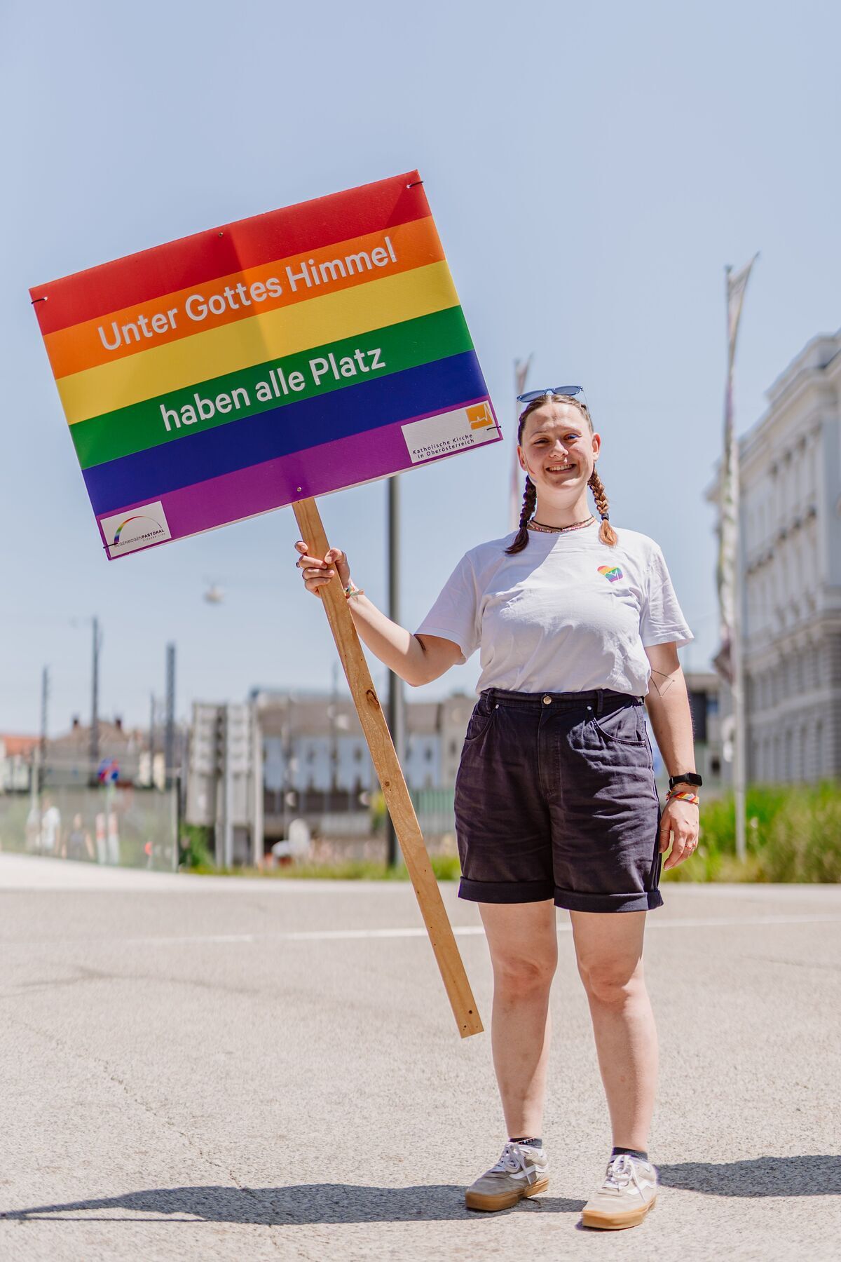 Agnes Schützenhofer, Jugendbeauftragte in der Pfarre Tassilo-Kremsmünster und ehrenamtlich engagiert in der Regenbogenpastoral, trägt das Schild 'Unter Gottes Himmel haben alle Platz'.