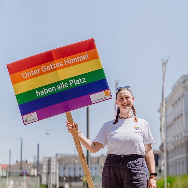 Agnes Schützenhofer, Jugendbeauftragte in der Pfarre Tassilo-Kremsmünster und ehrenamtlich engagiert in der Regenbogenpastoral, trägt das Schild 'Unter Gottes Himmel haben alle Platz'.